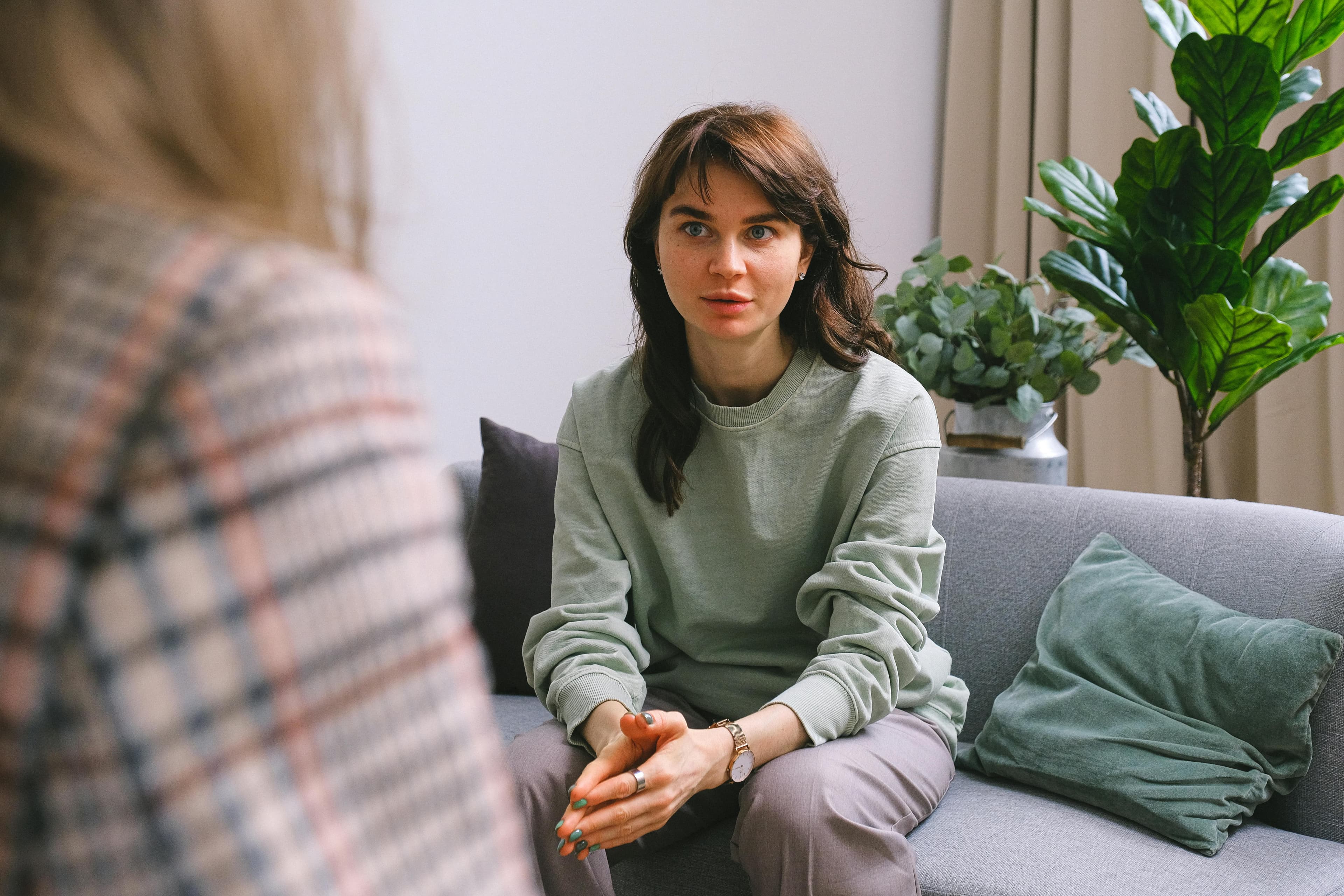 En kvinne sitter i en grå sofa. Hun har mørkt langt hår, lysegrønn genser og lyse bukser. Hun snakker til en annen person som har på seg en rutete dressjakke.
