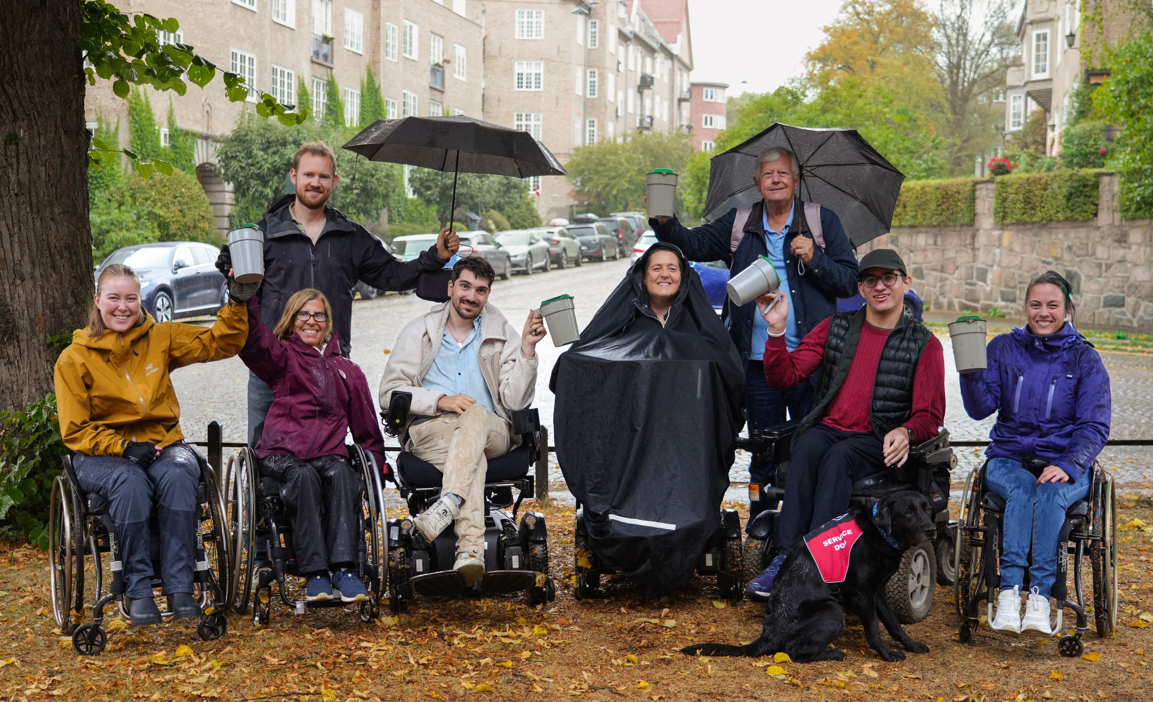 Flere mennesker står utendørs, noen sitter i rullestol og andre stå. Noen bruker også paraply. Alle holder i hver sin bøsse i forbindelse med innsamling til TV-aksjonen 2025