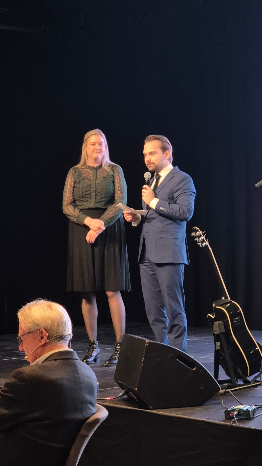 To personer står oppe på en scene og holder en tale ut til en større forsamling. Kvinnen har på seg et mørkt skjørt og en grønn topp. Hun har lyst hår. Mannen har på eg en blå dress. Han har mørkt hår.