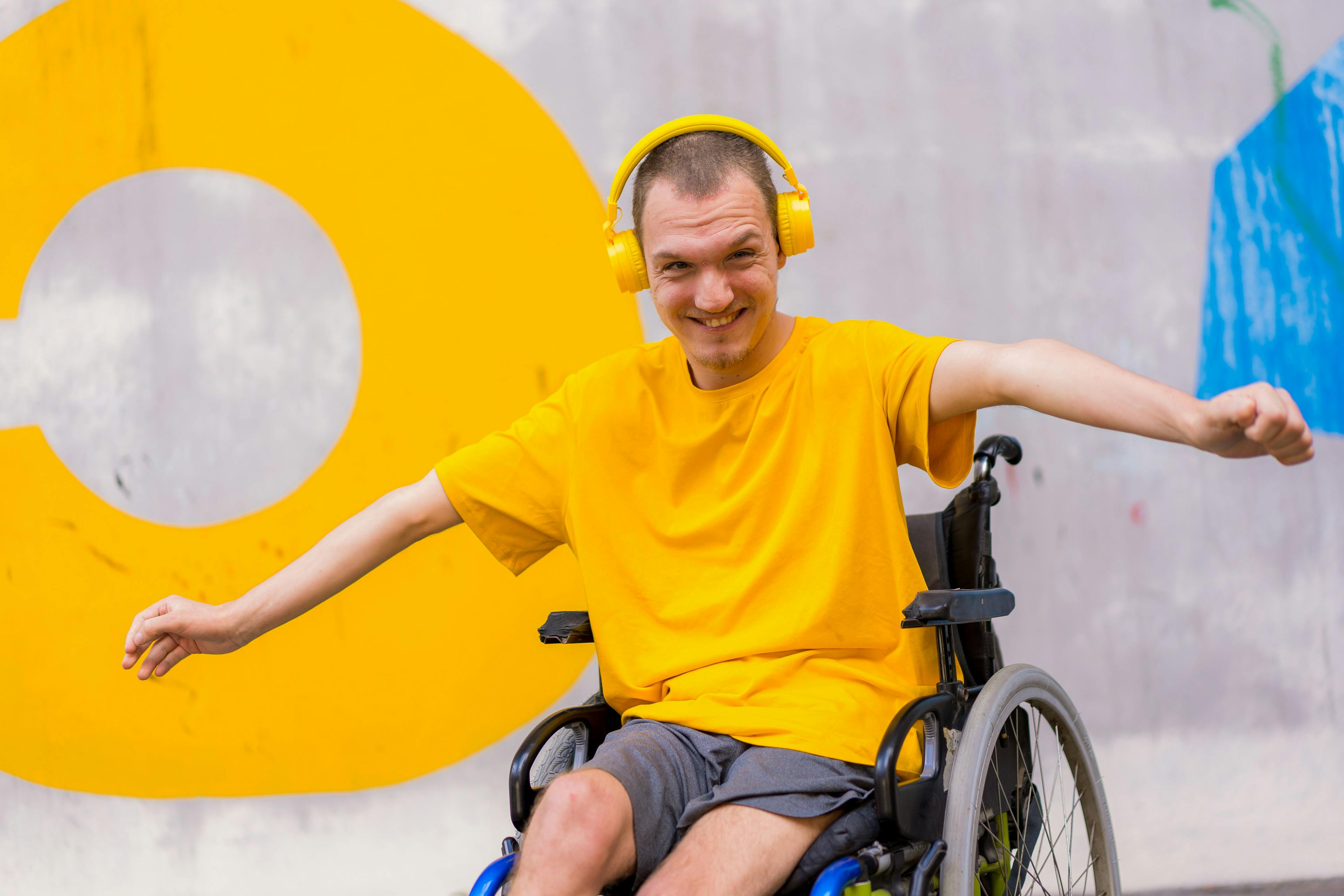 En mann smiler bredt mot kamera. Han sitter i rullestol og har på seg en grå shorts og gul t-skjorte. På hodet har han et gult headsett.