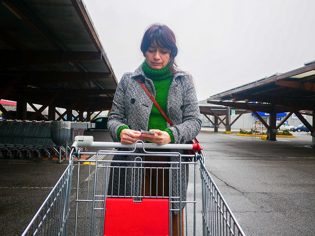 Kvinne med en handlevogn står på en stor parkeringsplass utenfor et kjøpesenter. Hun ser bekymret på handlelappen.