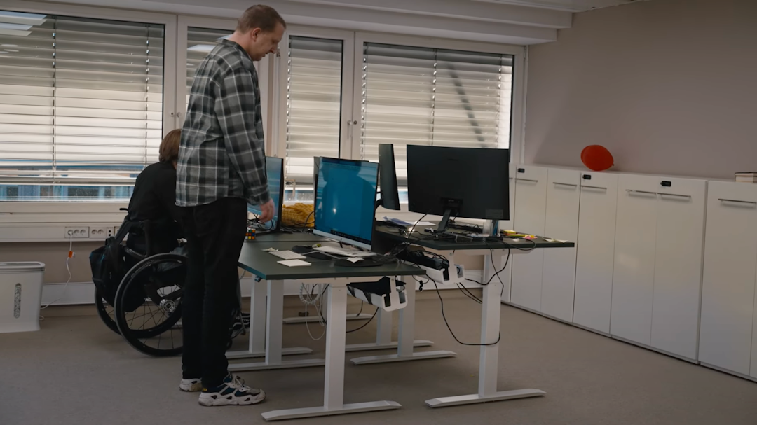 I en kontorlandskap står en høy mann foran et skrivebord med en datamaskin. Mannen har ingen stol å sitte på. Ved siden av er en person i rullestol som sitter å jobber.
