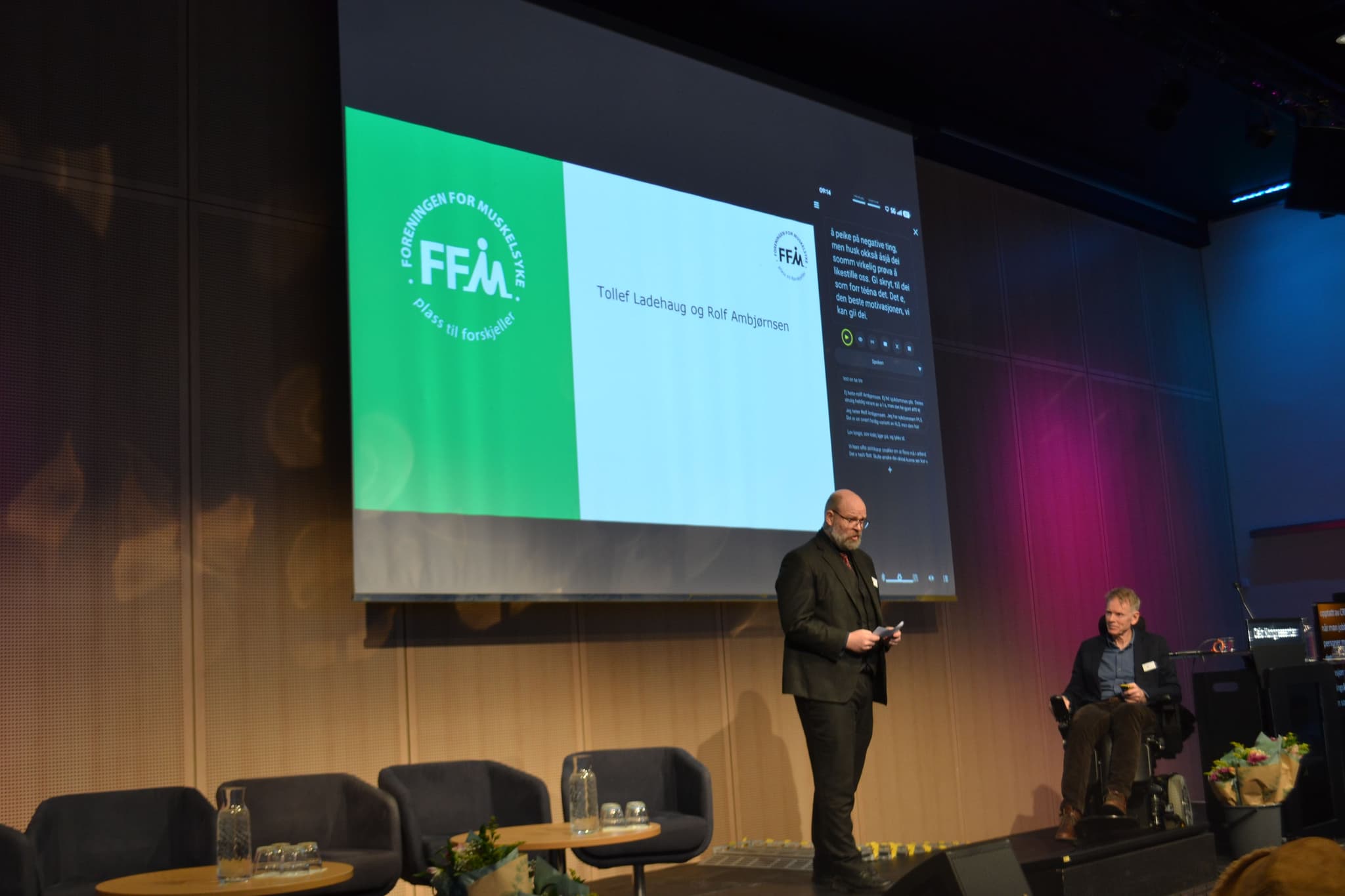 To personer står oppe på en scene. De har på seg dress. En sitter i rullestol. Bak dem er den en presentasjon.