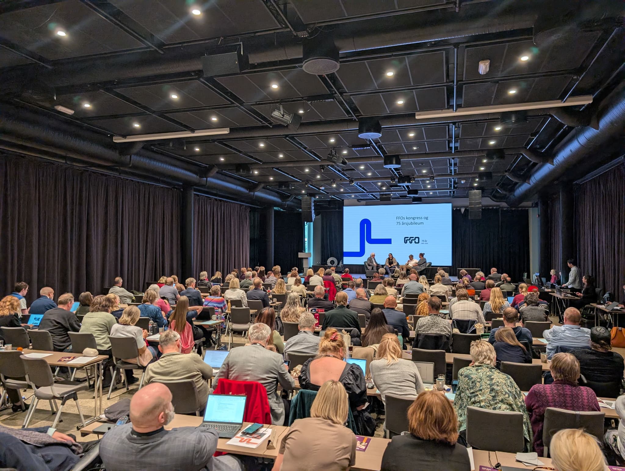 Mange personer sitter samlet i en stor sal. På en større scene sitter flere personer og fører en samtale. Bak dem er det en stor skjerm hvor det står: FFOs kongress og 75 årsjubileumsmarkering, med FFOs logo hvor det står 75 år og 1950-2025