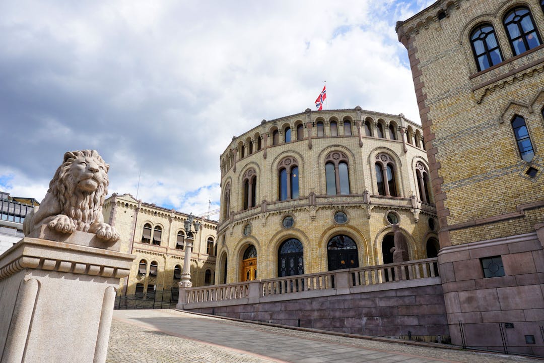 Stortinget i Oslo sentrum. Bygget har to firkantede bygg med et rundt et i midten. Mange vinduer og et norsk flagg vaier på toppen av den runde delen. Foran bygget står det en løve i stein som ligger.
