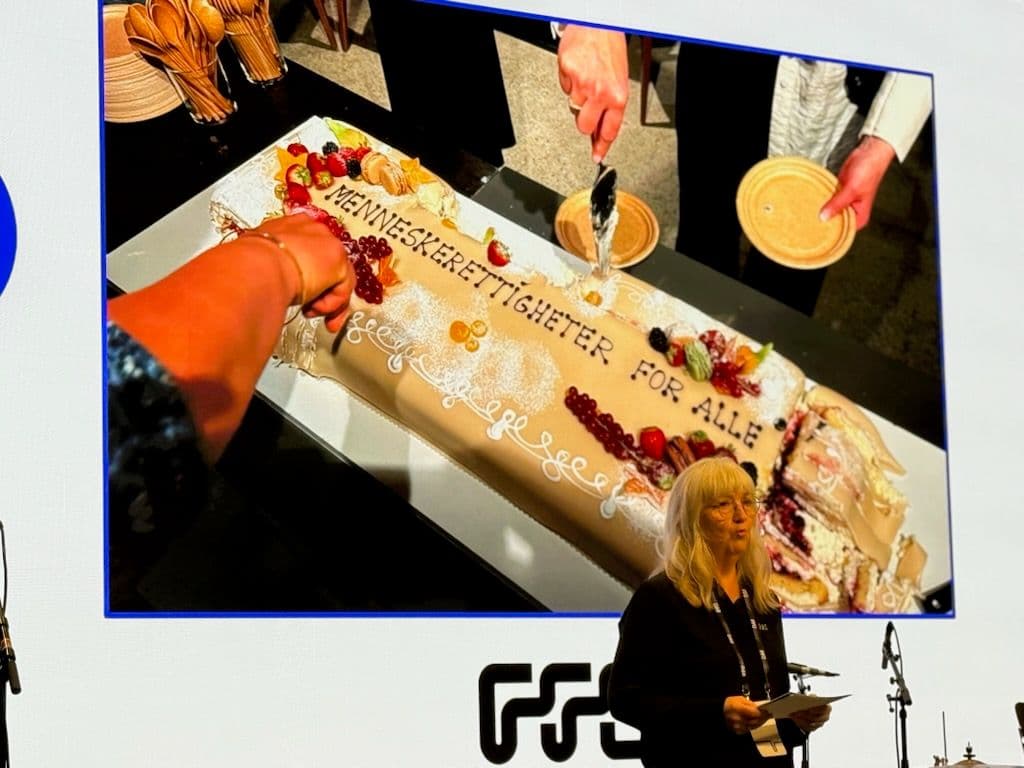 En kvinne står på en scene og holder en tale foran en større forsamling. Hun har på seg en mørk topp, har langt lyst hår og bruker briller. Bak hun er det bilde av en stor kake hvor det står: Menneskerettigheter for alle. Under står jubileumslogoen til FFO for 75 år.