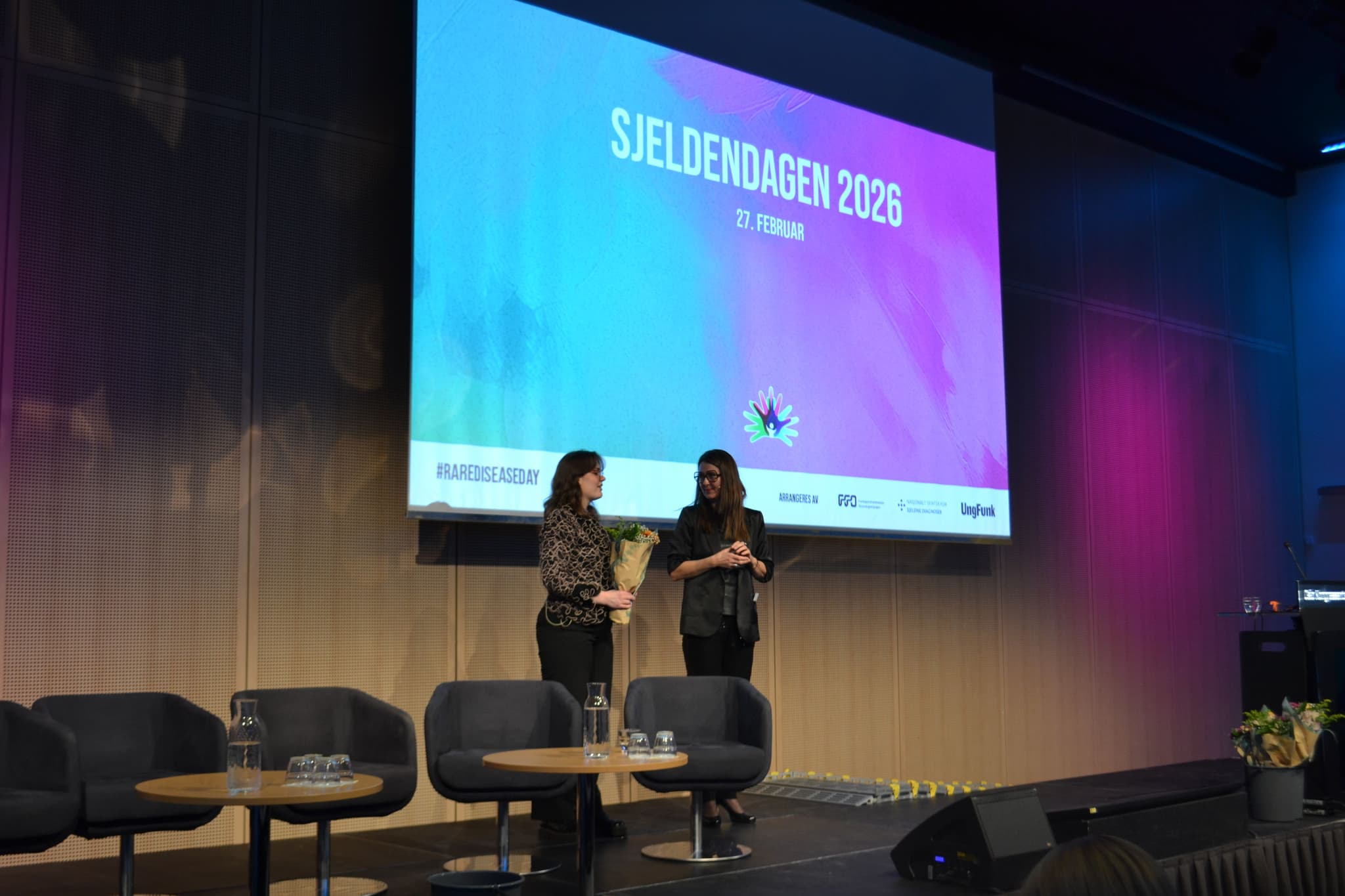 To personer står på en scene. Den ene personen blir takket av fra scenen med blomster.