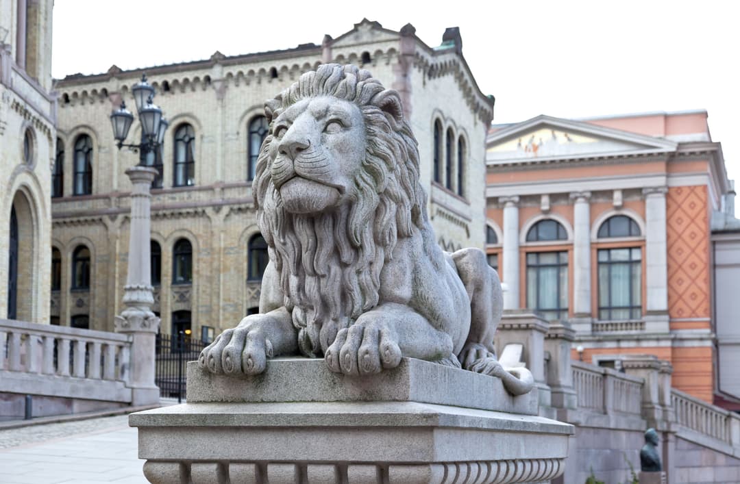En løve i stein som ligger på en blokk. I bakgrunnen er Stortinget i Oslo sentrum. Det er et bygg av lys stein og mange vinduer.