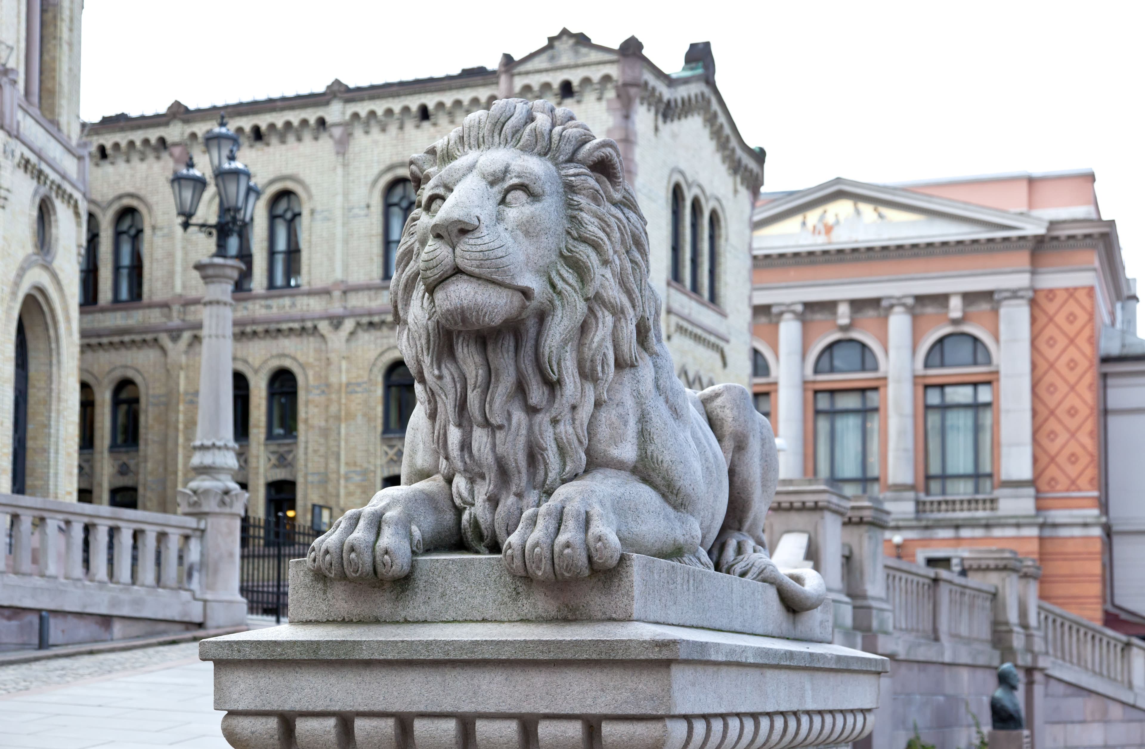 En løve i stein som ligger på en blokk. I bakgrunnen er Stortinget i Oslo sentrum. Det er et bygg av lys stein og mange vinduer.