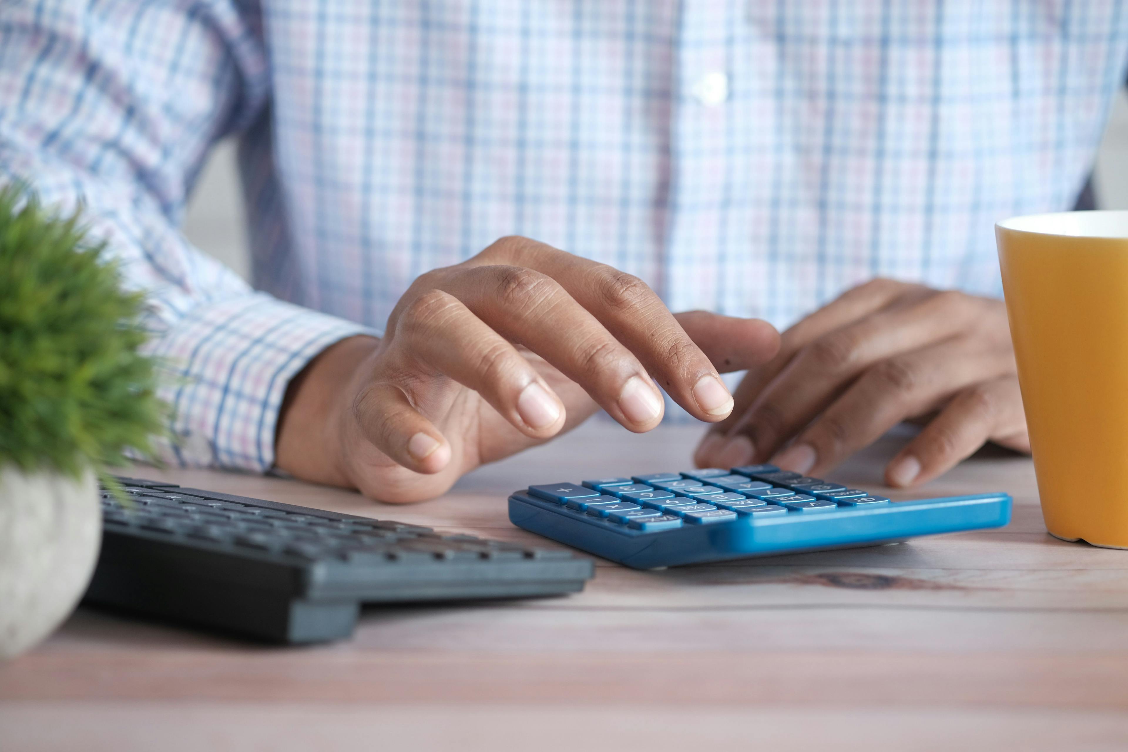 Hender som taster på en blå kalkulator. Til venstre ligger et trådløst tastatur og en grønn plante. Til høyre står det en gul kopp.