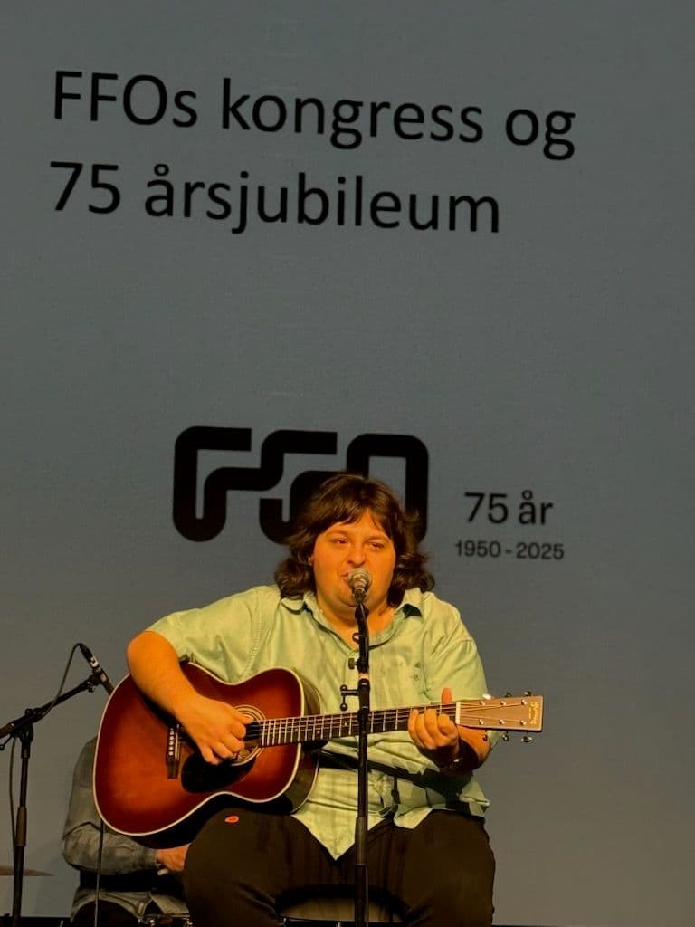 En ung gutt sitter på en scene. Han spiller på en brun kassegitar og synger inn i en mikrofon. Han har på seg mørk bukse og en turkis skjorte. Han har mørkt hår.