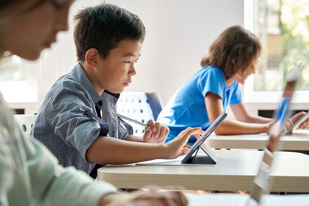 Tre elever på grunnskolen sitter i et klasserom. I fokus er det en elev i rutete skjorte som sitter å trykker på et nettbrett.
